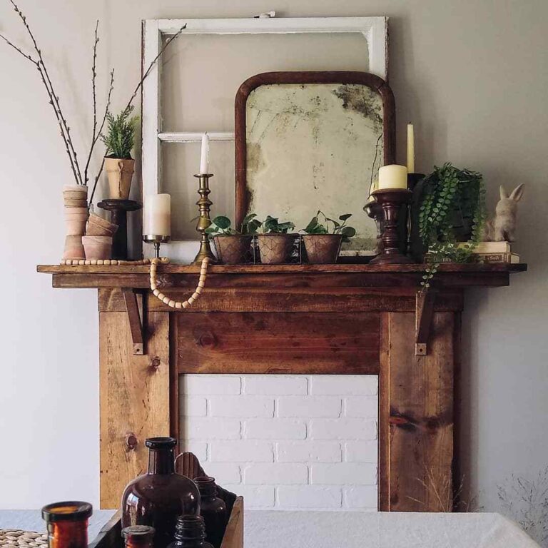 A vintage wooden mantel with white bricks in place of the fireplace, showcasing a wooden white window frame, old mirror, a brass candlestick holder with a white candlestick, small clay flowerpots, and other vintage items.