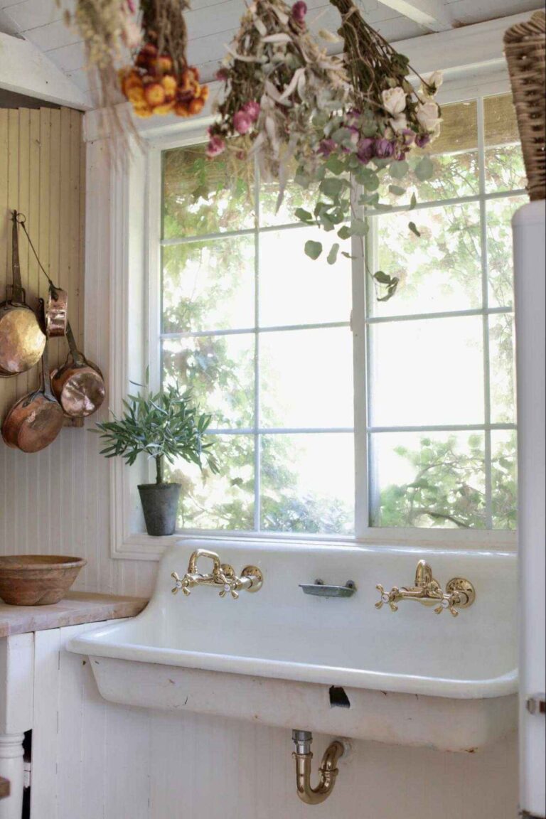 Part of a country cottage kitchen with a vintage farmhouse sink with two gold-tone faucets and a window above it. Dried flowers hang above the sink and copper pans to the left.