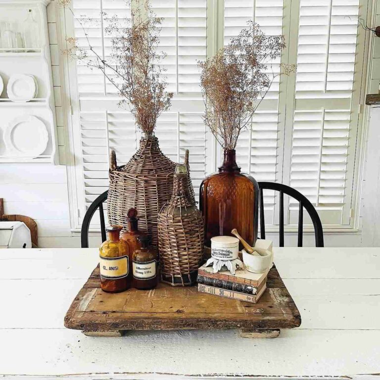 A white rustic wooden table displaying a vignette made up of an old wooden tray with amber bottles, wicker bottle containers, and vintage books.