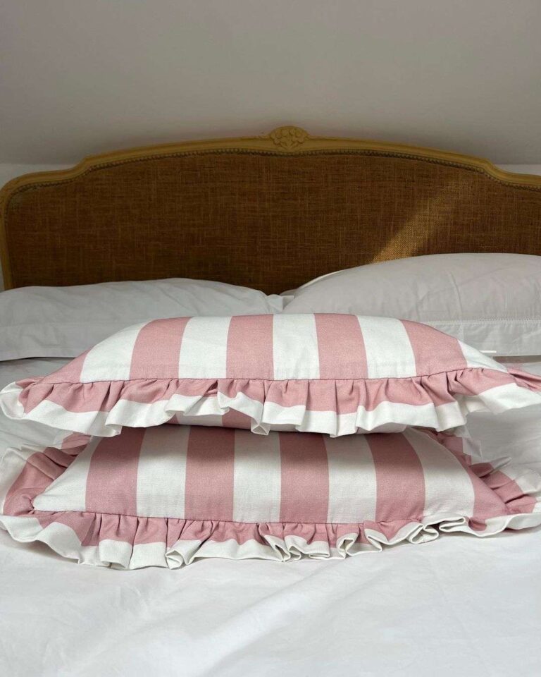 Two rose pink and white striped pillows with ruffled edges stacked one on top of the other, sitting on white bed sheets.