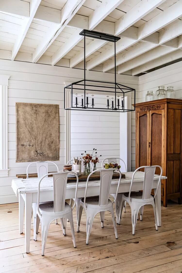 A simple wooden white table with six white metal chairs stands on a light brown wooden paneled floor. White shiplap walls are seen in the background with antique tapestry, an industrial hanging lamp hangs from the ceiling, and a vintage wooden wardrobe is seen to the right.