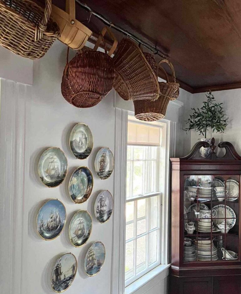 Nine round vintage plates displaying old images of ships hang on a white wall, next to them a window, and an antique dark wood cabinet with vintage white and blue dinnerware inside. Wicker baskets hang from a wooden ceiling.