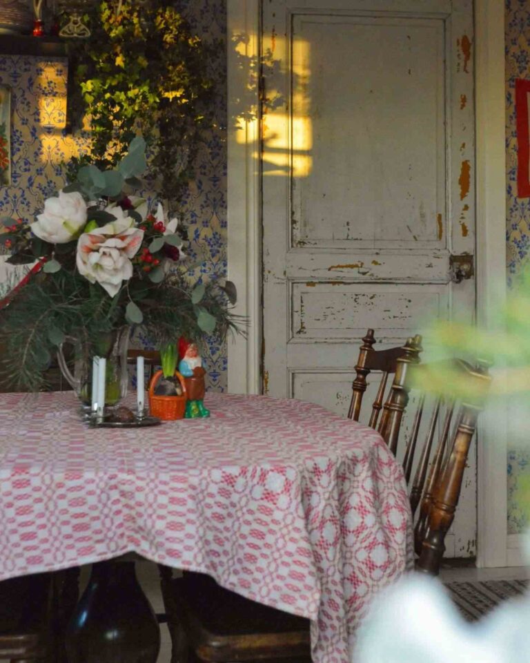 Afternoon light in a vintage Scandi farmhouse featuring a partial view of an oval table with a red and white patterned tablecloth, two vintage wooden farmhouse chairs, a floral decoration on the table, vintage blue and white patterned wallpaper, and a worn-out shabby rustic white door.