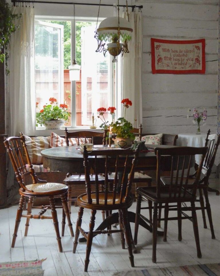 The interior of a vintage farmhouse in the Scandinavian style featuring a vintage oval wooden table with vintage wooden farmhouse chairs, a white wood paneled floor, a window with white curtains, flowerpots of red geraniums, an antique hurricane lamp hanging from the ceiling, and a small old red and white tapestry on a white rustic wooden wall.