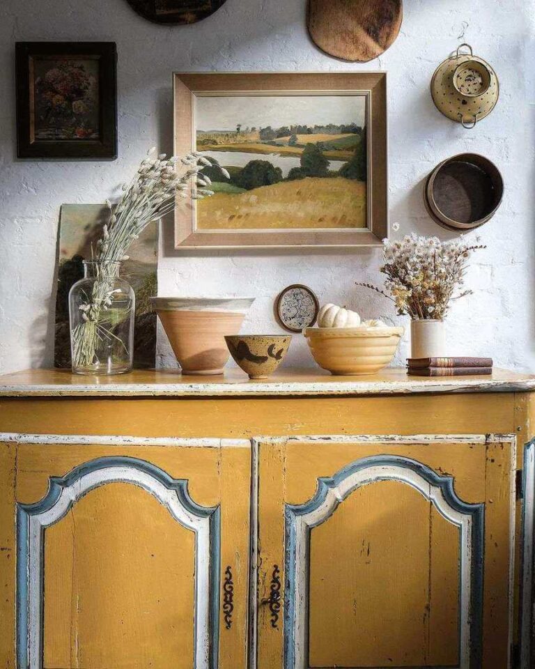 A vintage reclaimed furniture yellow floor cabinet with white and dark blue finishes with vintage vessels, flowerpots, and books on top. An antique painting of a pastoral scene and some vintage decorations hang on the white wall.