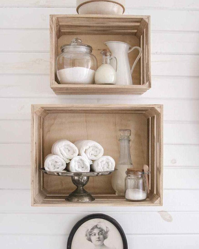 White horizontal paneled wall with two wooden crates used as shelves displaying white hand towels on a vintage pewter tray and glass containers with bath and body products. Part of a black oval frame with a black and white photo is seen below the bottom crate.