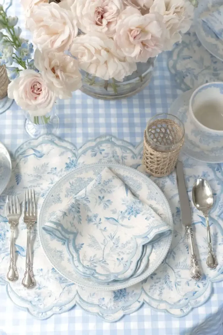 A vintage blue and white table setting featuring a light blue gingham tablecloth, a blue and white English transferware plate, vintage floral blue and white napkins, antique silverware, and pale pink roses.