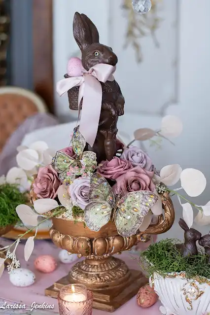 An antique-style Easter tablescape with a gilded planter, pink and purple roses, and a chocolate bunny wearing a pink satin ribbon bow around his neck.