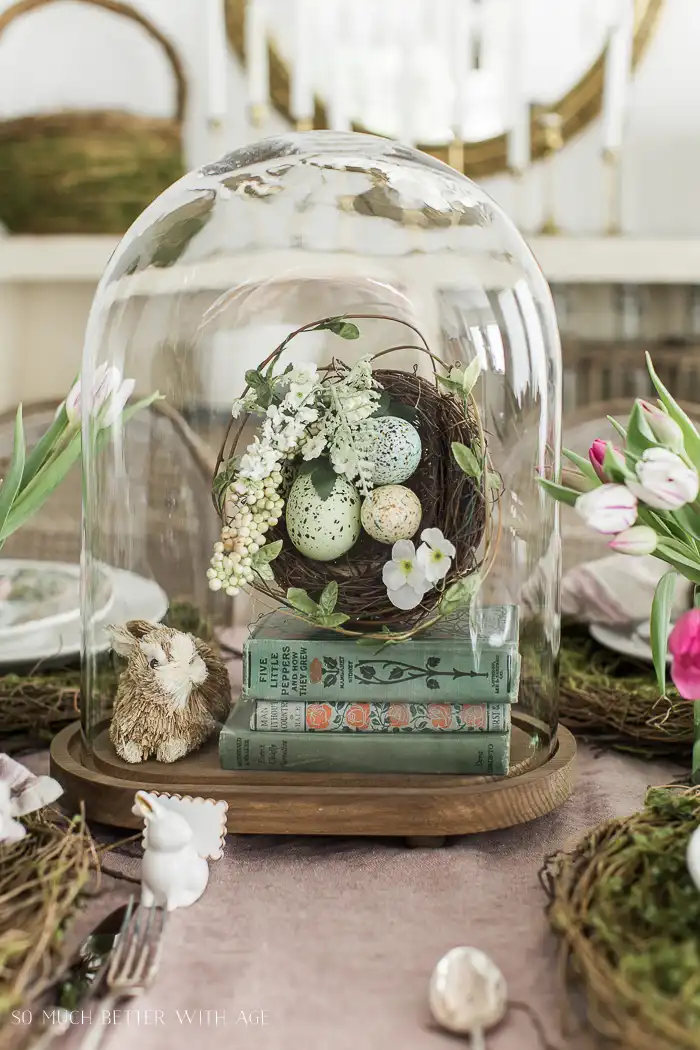 A vintage Easter tablescape in a bell jar featuring three green vintage books, a vintage bunny figurine, and a flower-decorated nest with quail eggs.