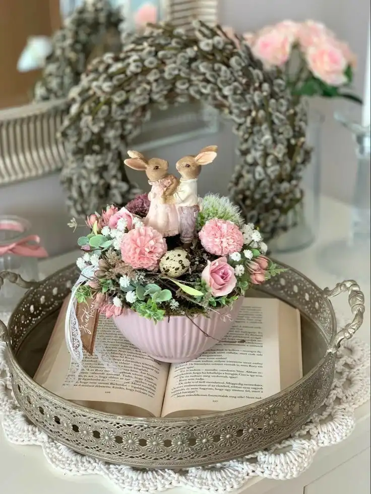 A vintage Easter teacup decoration in a light pink bowl, featuring faux plants, quail eggs, a lace ribbon, and two figurines of newlywed bunnies, sits on a round vintage tray.