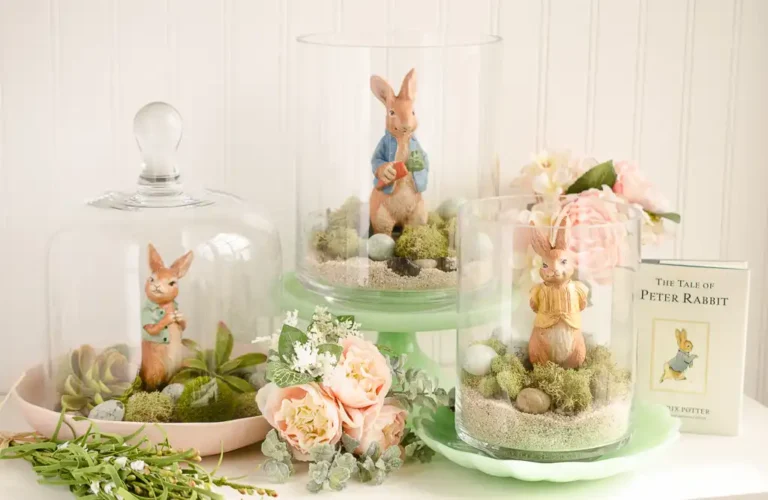 A vintage Easter vignette featuring a glass cloche and two open glass containers with a vintage figurine from The Tale of Peter Rabbit in each, with other floral and egg decorations.