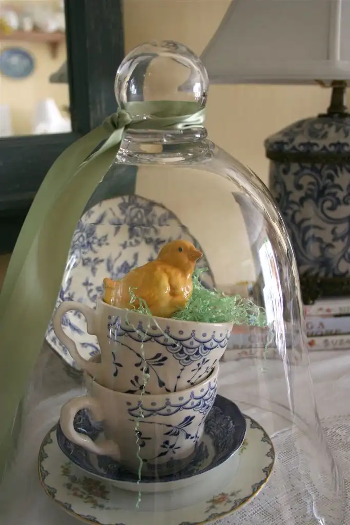 A vintage Easter vignette inside a glass cloche featuring vintage blue and white teacups and a vintage chick figurine.