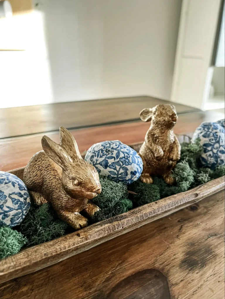 Two vintage gold bunny figurines and three decoupaged eggs with blue and white floral motifs sit on moss in a wooden container.