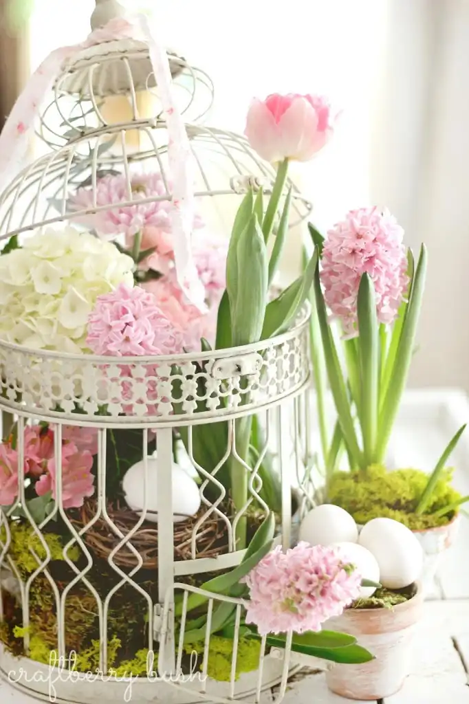 A white romantic vintage birdcage displaying a vintage Easter vignette made up of pink hyacinths, light green moss, a pink tulip, and a small brown twig nest with a white egg. A small terracotta flowerpot with moss and three white eggs and another terracotta flowerpot with moss and a pink hyacinth stand next to the cage.