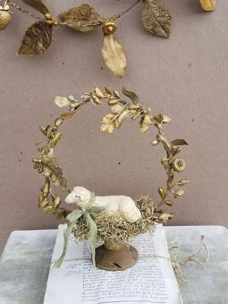 A vintage Easter centerpiece made from a gold spray-painted metal circle with floral ornaments and a vintage sheep figurine sits on an old vintage book.