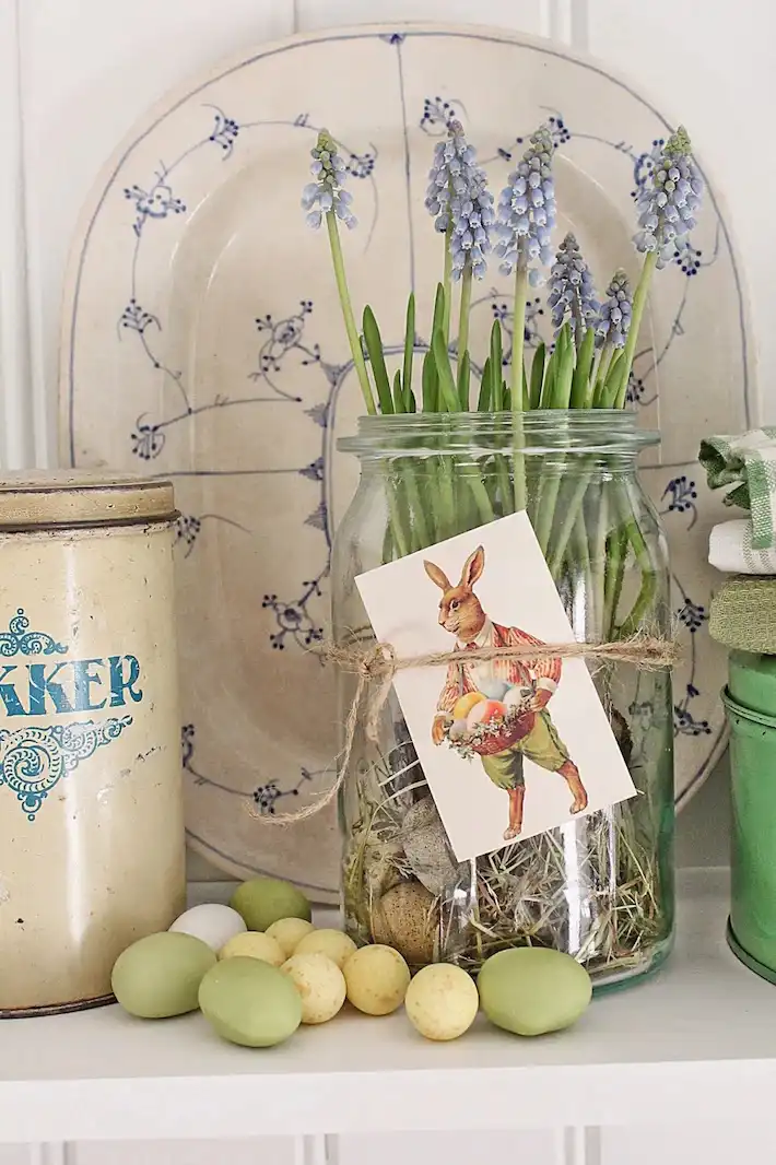 A glass jar with grape hyacinths and a vintage Easter postcard tied around it using twine. Pastel green eggs are spread in front of the jar and a vintage blue and white platter is seen behind.