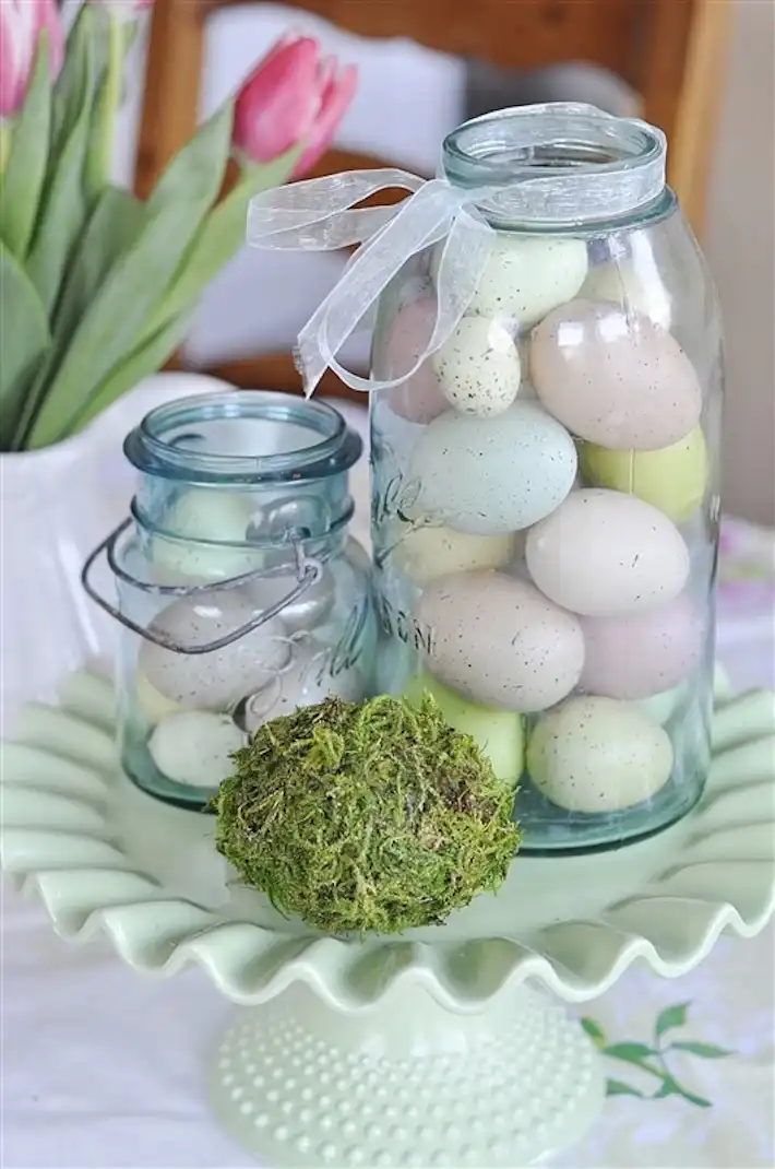 Two Mason jars filled with pastel-colored eggs stand on a vintage jade-colored cake stand.