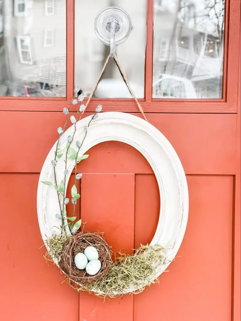 A vintage Easter wreath made from an old oval white frame with an egg nest, dried moss, and faux pussywillow branches hangs on a vintage orange door.