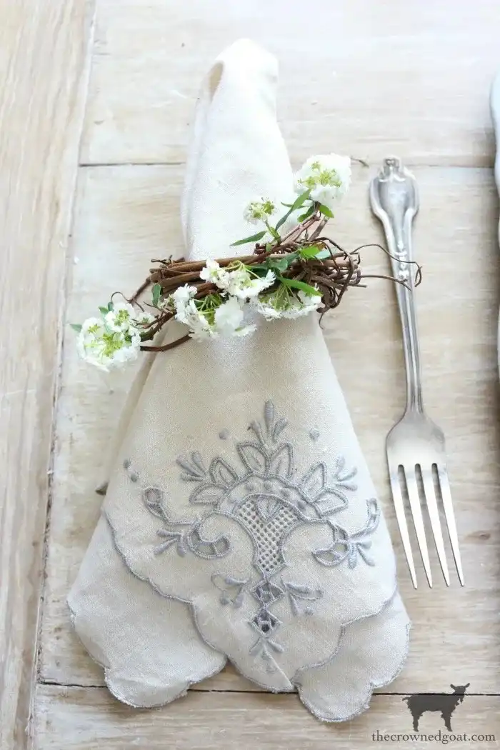 A nicely folded light blue and white linen napkin with decorative motifs with a napkin ring made out of twigs and white flowers.