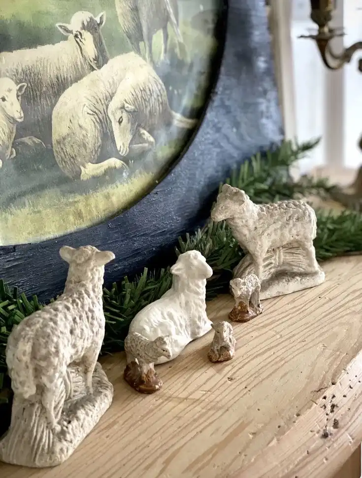 Three big and three small vintage sheep figurines stand on a wooden rustic surface with an old painting of sheep in a black vintage frame behind them.