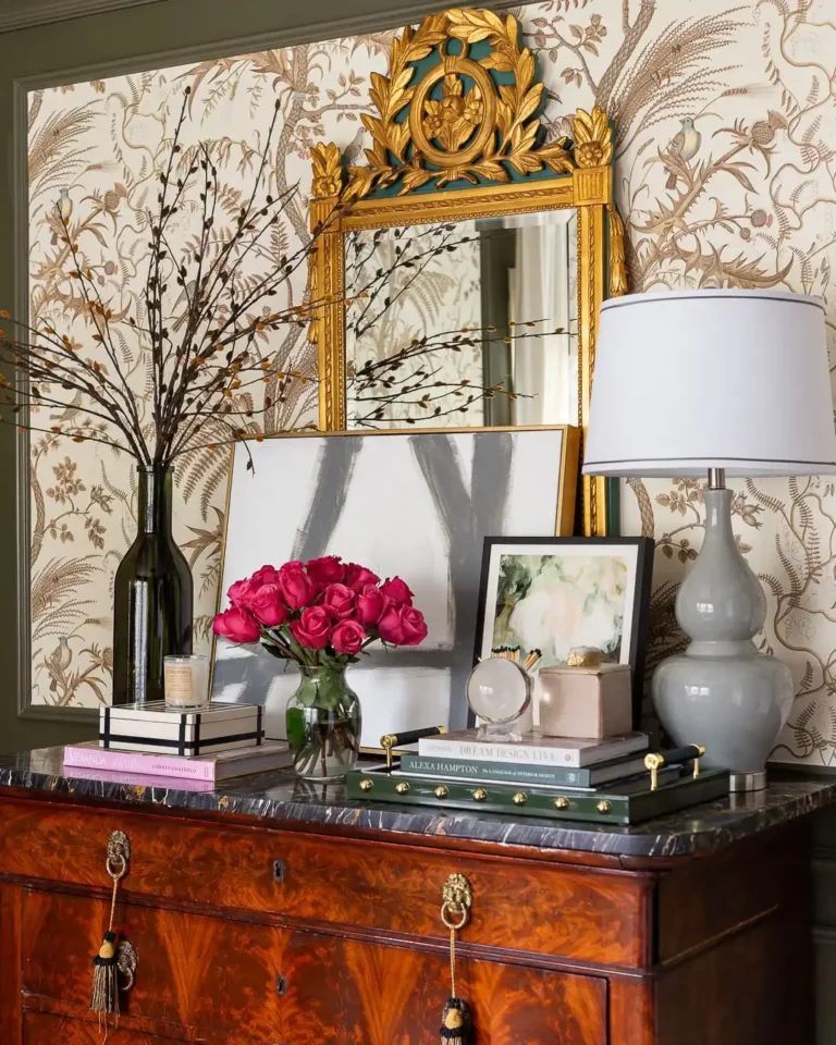 An Art Deco-inspired vignette made up of coffee table books, a bouquet of pink roses, a vintage lamp, an abstract painting, and an ornate gold-frame mirror on an old Hollywood glamour dresser.