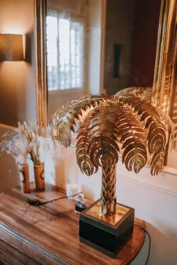 A gold-tone palm tree decoration stands on a wooden surface with a rectangular mirror in a gold frame seen in the background.