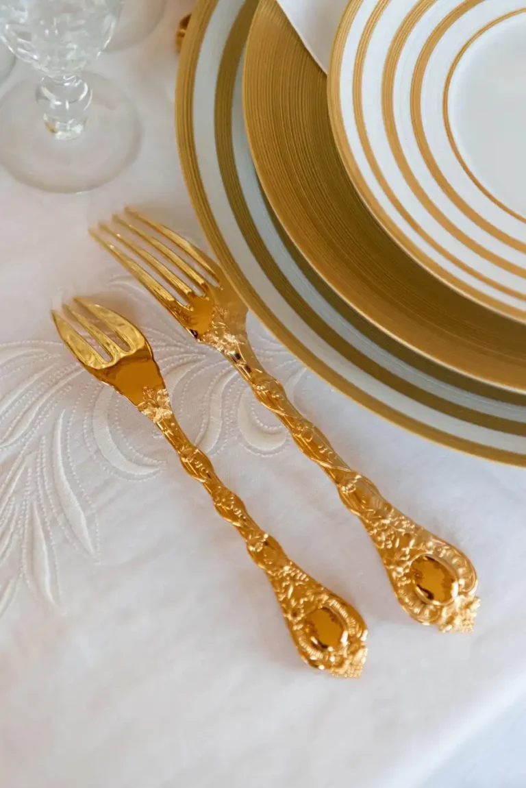 One small and one large gilded fork on a floral white tablecloth next to a white and gold plate set.