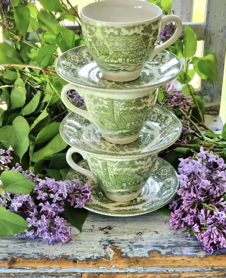 Three vintage green English transferware teacups on plates stacked on top of one another, sitting on a rustic washed wooden surface with lilac flowers on either side.