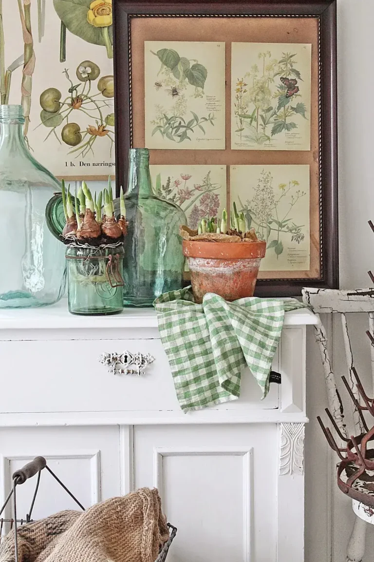 Vintage green bottles, a vintage green jar filled with Spring flower bulbs, a vintage clay pot with sprouting Spring flowers, and antique botanical prints stand on a rustic white piece of furniture.