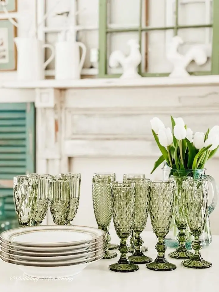 Twelve vintage green glasses on a white surface, five white soup bowls stacked on top of one another, and a bouquet of tulips in a glass jug.