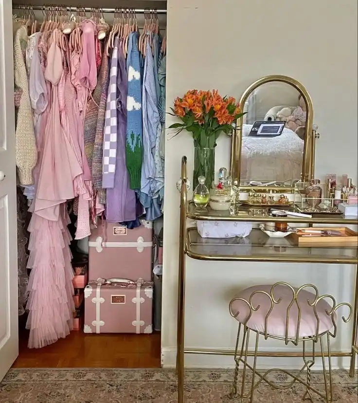 An open closet with pink and purple clothes and two pink and white suitcases; next to it a gilded vintage Hollywood glam vanity table with perfume and makeup accessories.