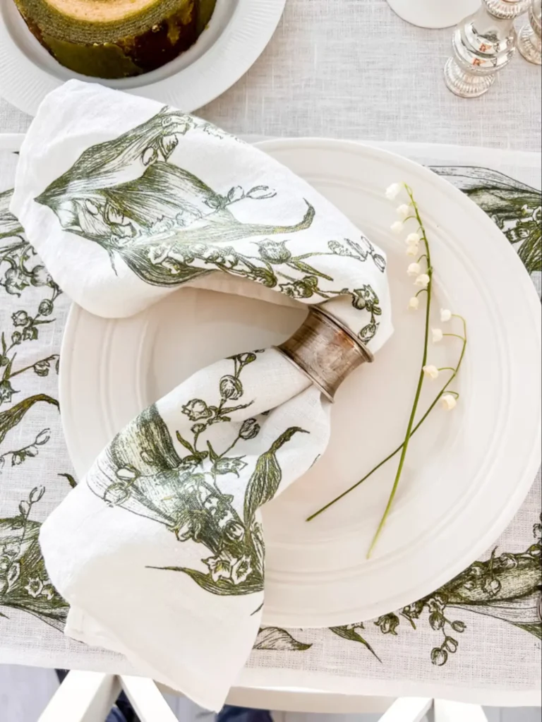 A vintage linen napkin with a dark green illustration of lily of the valley sits on a white plate on a vintage Spring decor table.