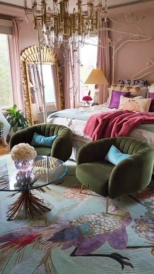 A room with two scalloped green velvet armchairs, a colorful vintage rug with a bird and flower print, a big rectangular floor mirror with a gold ornate frame, and a bed with colorful pillows in the background.