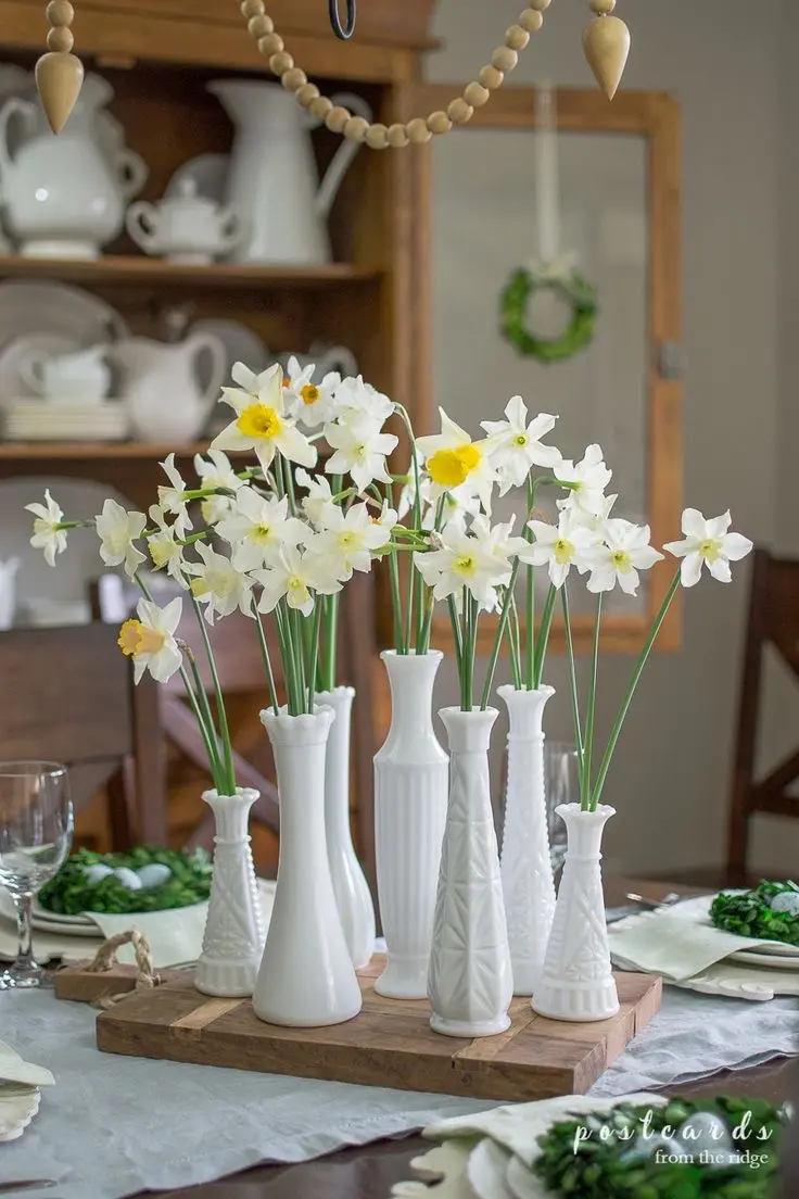 Seven milk glass vases of various sizes with narcissus flowers sit on a wooden cutting board in a vintage Spring decor table setting.