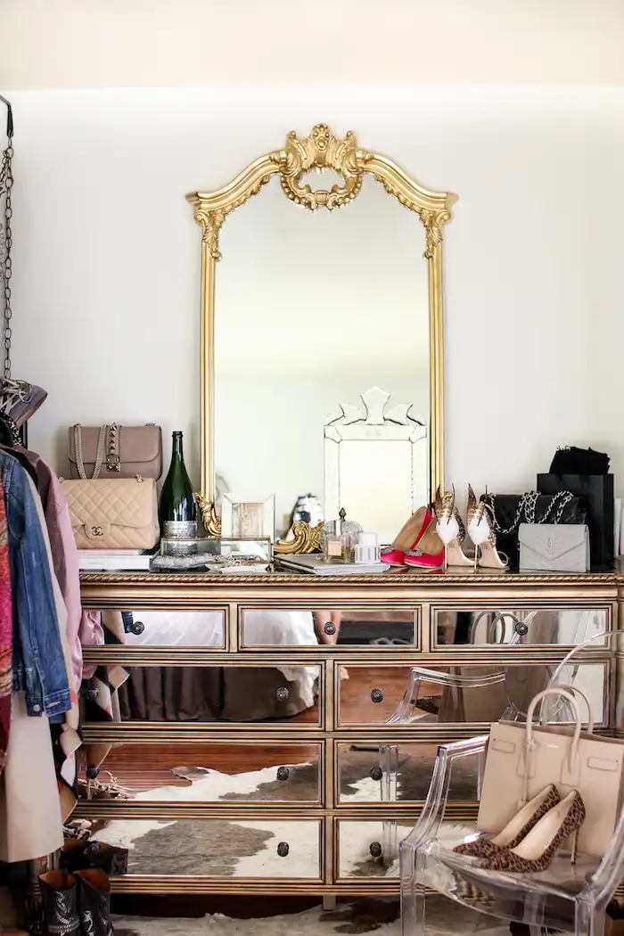 A vintage mirrored dresser with a bottle of champagne, Chanel handbags, stiletto heels, perfume, and an ornate gilded mirror. A plastic chair with leopard print stiletto heels and a beige handbag stands to the right.