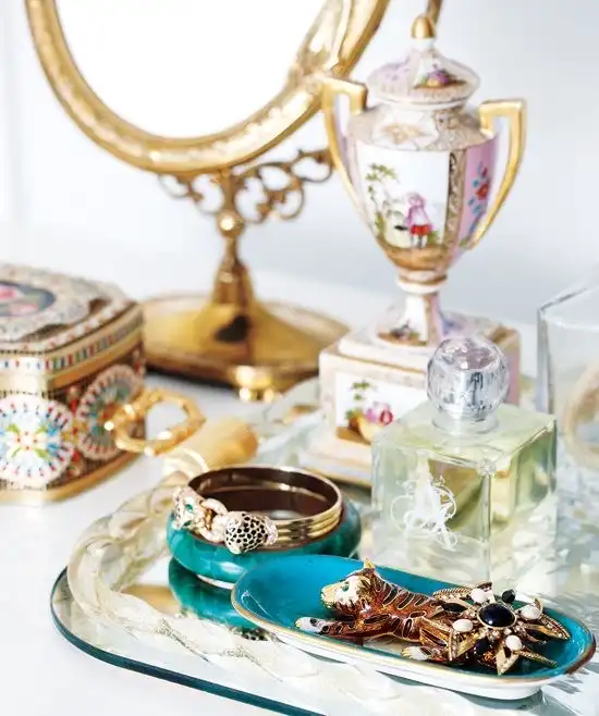 A closeup view of old Hollywood glam accessories on a table, including a decorative pink and gold container, an oval gold frame makeup mirror, and jewelry and a perfume bottle on a mirrored tray.