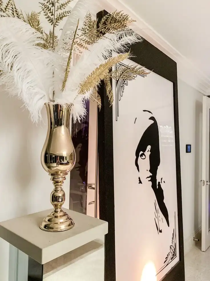 A bouquet of white ostrich feathers in a tall golden vase stands on top of a small square shaped beige tabletop next to a huge black and white old-fashioned print of a woman.