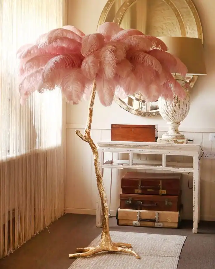 A pink ostrich feather floor lamp on a gilded leg stands in front of a rustic white side table; under the table there is a stack of vintage suitcases and on top a rustic white lamp with a vintage oval mirror behind it.