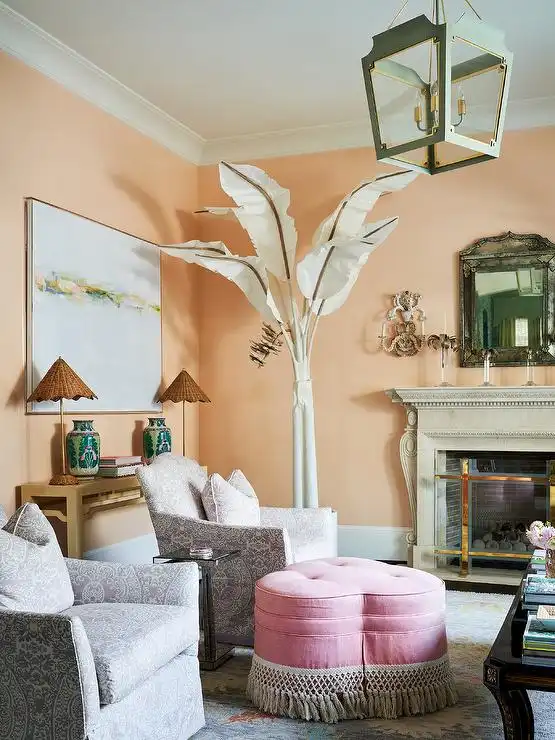 A faux white palm tree decoration in a room with peach-colored walls, two gray patterned armchairs, a pink heart-shaped ottoman with tassels, an antique fireplace, and a vintage hanging lamp.