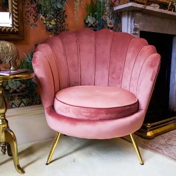 A pink velvet scalloped armchair on gold legs with a partially visible mantel and a piece of an orange wall with plants on it.