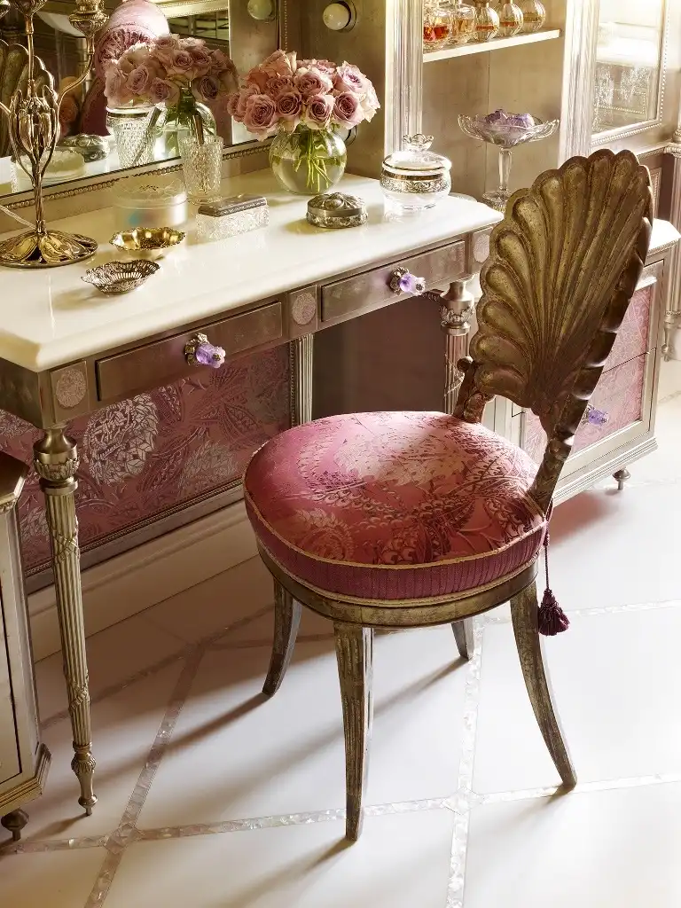 An antique vanity table with light purple crystal knobs, a gilded decorative chair with a seashell backrest, a bouquet of roses in a round green vase and makeup and jewelry accessories sit on the vanity table.