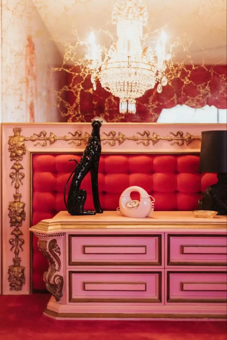 Anornamtental old Hollywood glam dresser in pink and gold with a black statue of a cat stand against a red tufted velvet wall encased in a white frame with gold ornaments, and a mirror behind it.