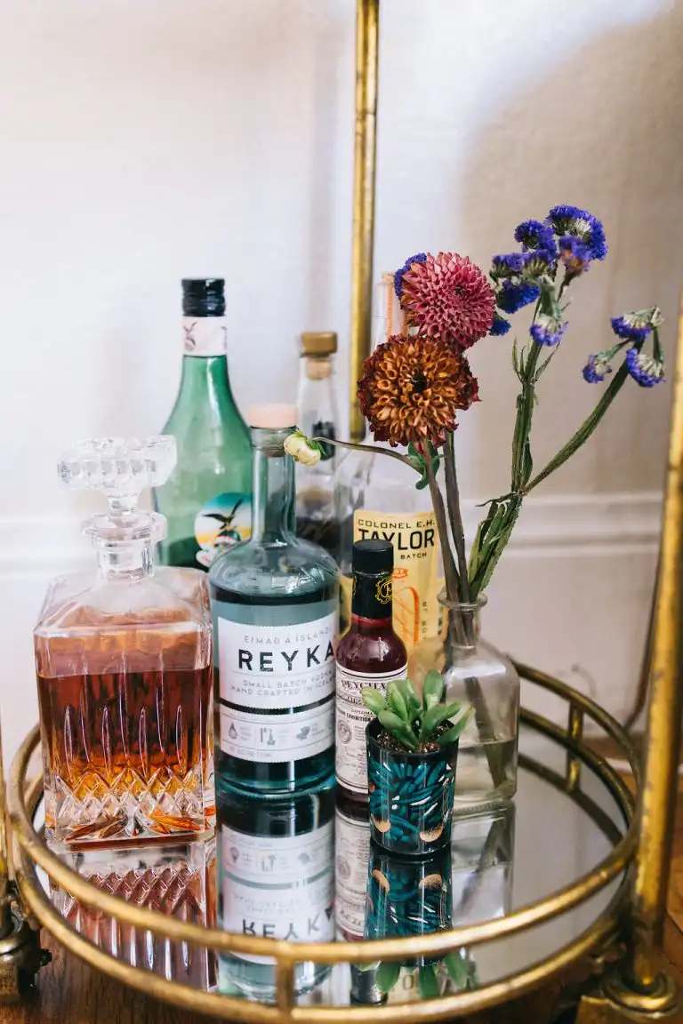 A round gilded bar cart with a mirrored surface, a couple of alcohol bottles, a crystal decanter, and a bouquet of wildflowers on top.