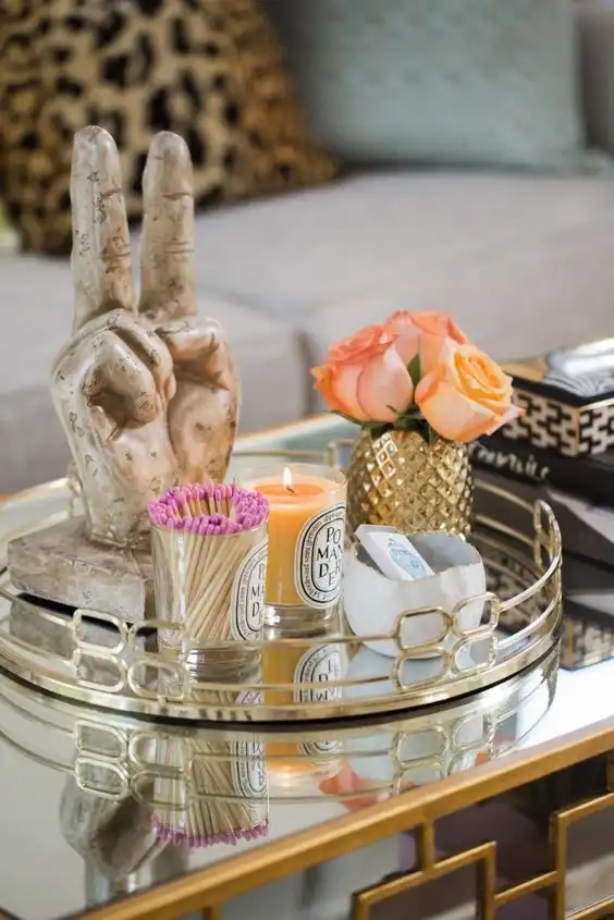 A Hollywood glam mirrored table with gold metalwork and a round mirrored tray with candles, a decorative hand, and a small bouquet of roses in a gilded vase.