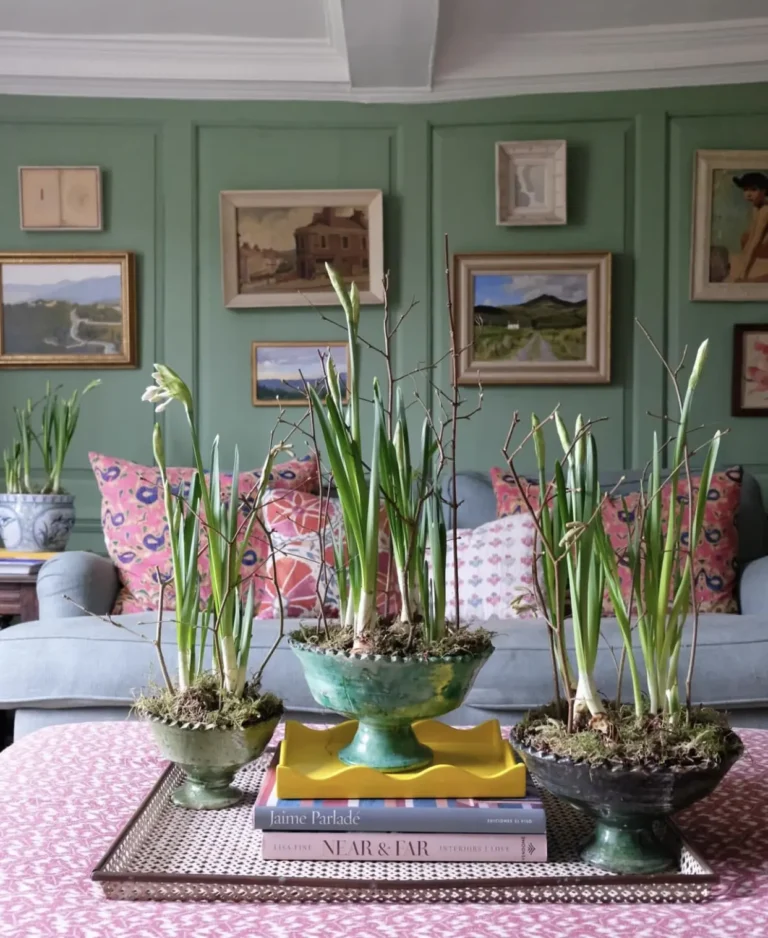 A vintage Spring home decor coffee table vignette featuring three vintage planters with snowdrops, books, a vintage tray, and a pink and white tablecloth. A couch with patterned throw pillows and a green wall with frames is seen in the background.