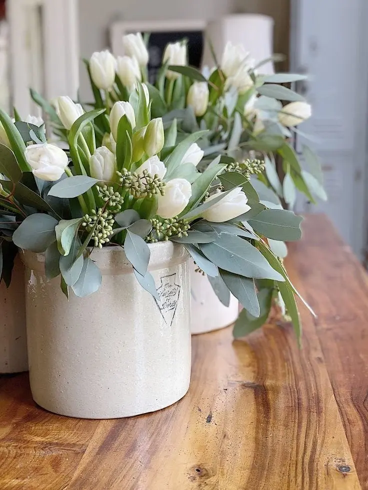 Bouquets of white tulips in cream-colored vintage crocks sit on a light wooden surface.