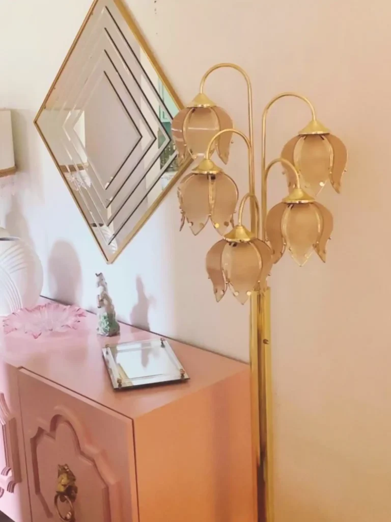 A vintage pink dresser with a mirrored tray and other Hollywood Regency decorations, a diamond-shaped gold-framed mirror, and a pink vintage floor lotus lamp.