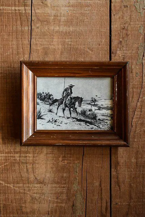 An old drawing in a rugged brown frame depicting a cowboy on a horse in a desert landscape with cactus plants in the background hangs on a rustic wooden brown wall.