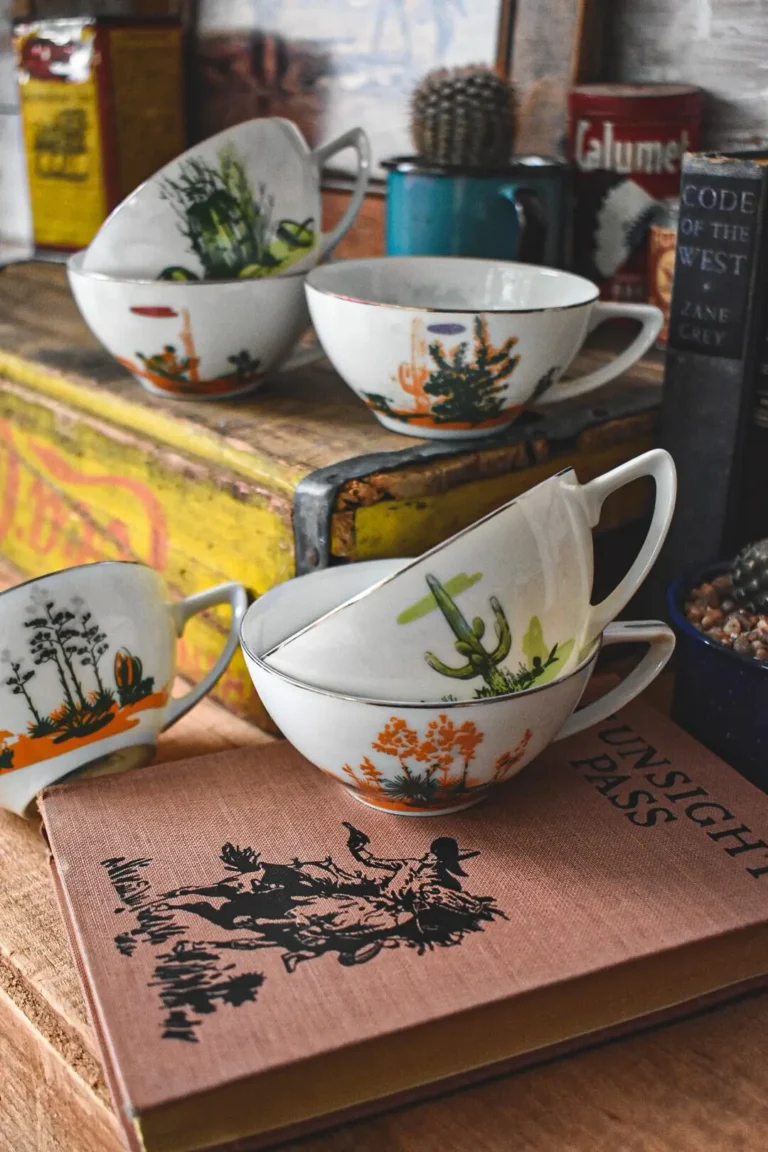 A couple of vintage teacups with cactus motifs displayed on top of an old western book and a yellow Coca-Cola crate.