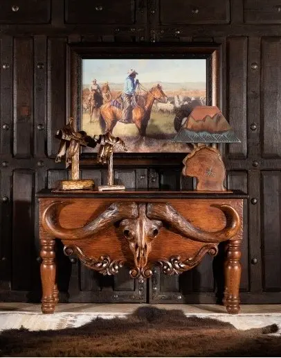 A cherry wood table with a carving of a big cow skull stands against a dark wood paneled wall with a Wild West-themed painting on it.